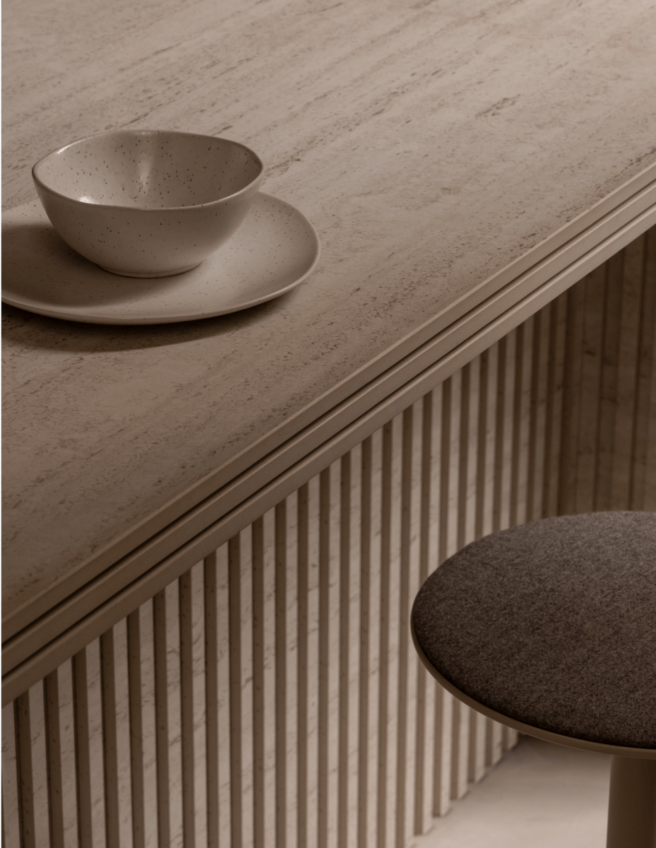 Modern minimalist interior with light beige sintered stone countertop, white ceramic bowl and saucer, vertical fluted paneling, and gray upholstered stool