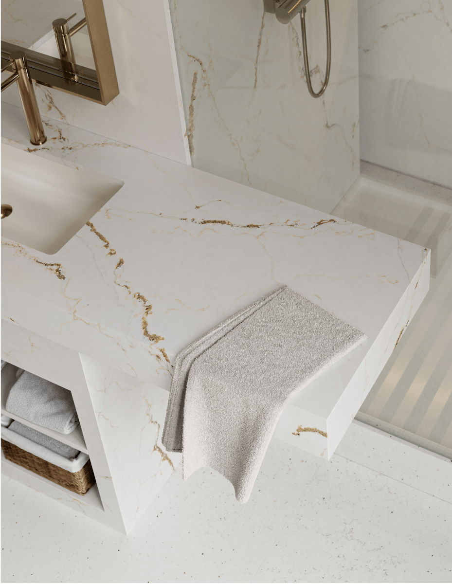 Modern bathroom vanity featuring white sintered stone countertop with elegant gold veining, integrated rectangular sink, brushed brass faucet, floating cabinet with open shelving holding folded towels and wicker basket, gray hand towel draped on counter edge, marble-look wall tiles, and frameless mirror reflecting shower area with rainfall showerhead