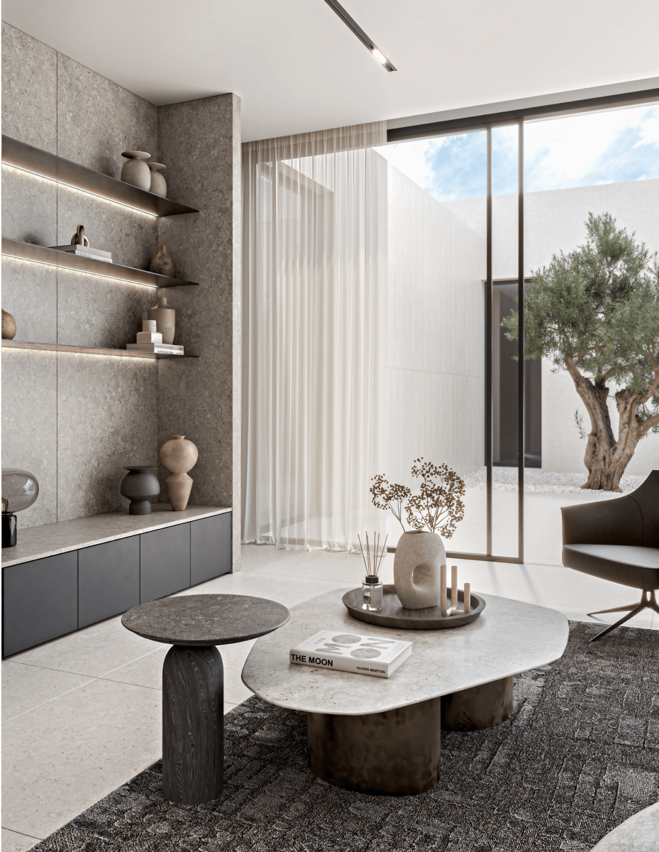 Contemporary living room featuring gray sintered stone accent wall with built-in illuminated shelving displaying ceramic vases and decor, oval marble coffee table with books and vase, modern armchair, floor-to-ceiling glass sliding doors opening to courtyard with olive tree, natural light, and minimalist interior design